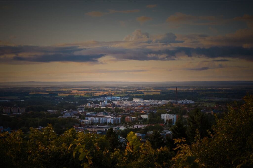 Utsikt över centrum med höghus, parkeringsplats, kyrktorn och Volvos fabrik sett från ovan i eftermiddagsljus. I bakgrunden breder milsvitt åkerlandskap och skog ut sig. / View over the city centre with high-rise buildings, car parks, church towers and Volvo’s factory seen from above in afternoon light. Sweeping expanse of field landscape and forest in the background.