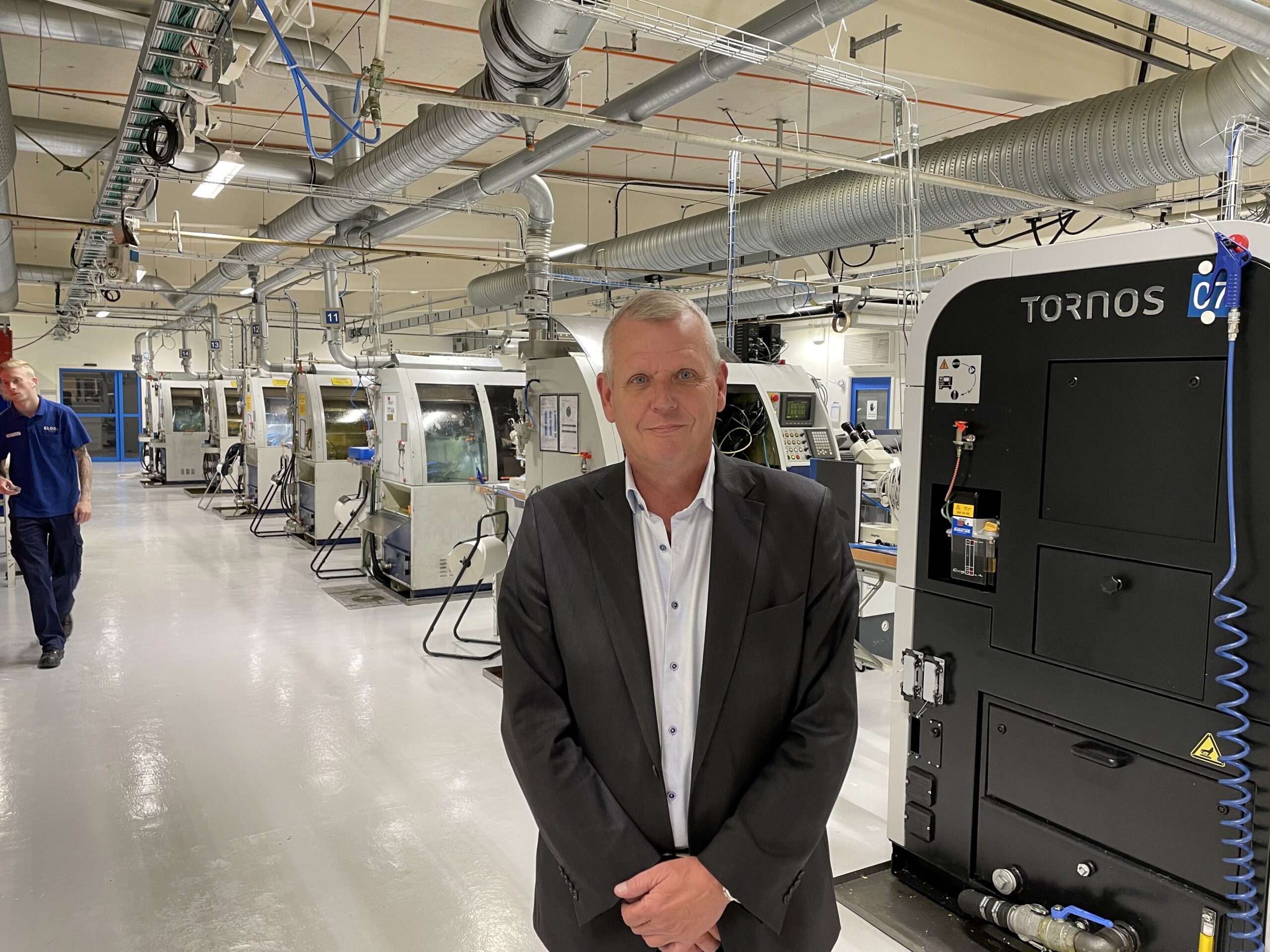 En man står i en fabrik med flera maskiner bakom sig.