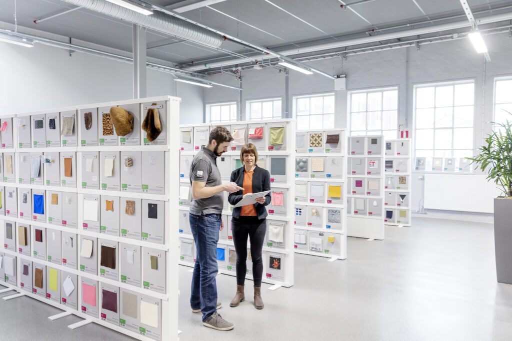 Two people stand in a show room with white shelves and look at a catalog of different materials.