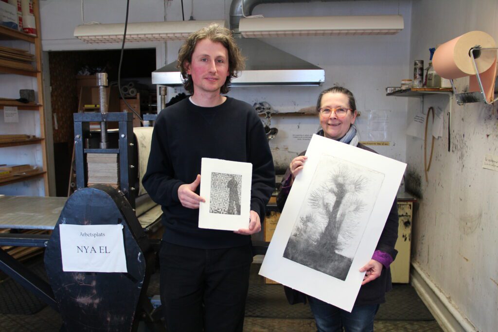 A man and a woman stand in a workroom with a printing press behind them. They each hold up a graphic sheet in front of them.