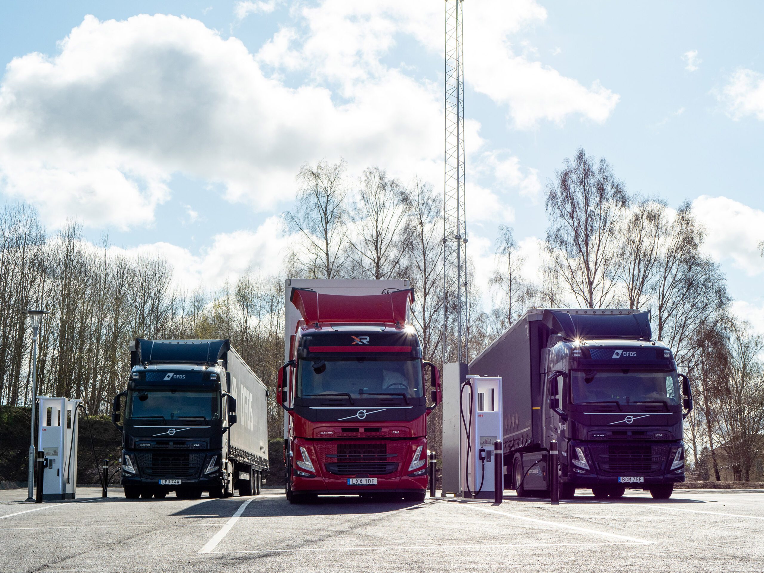 Three trucks are lined up with their fronts facing forward and between them are charging stations for heavy battery charging.