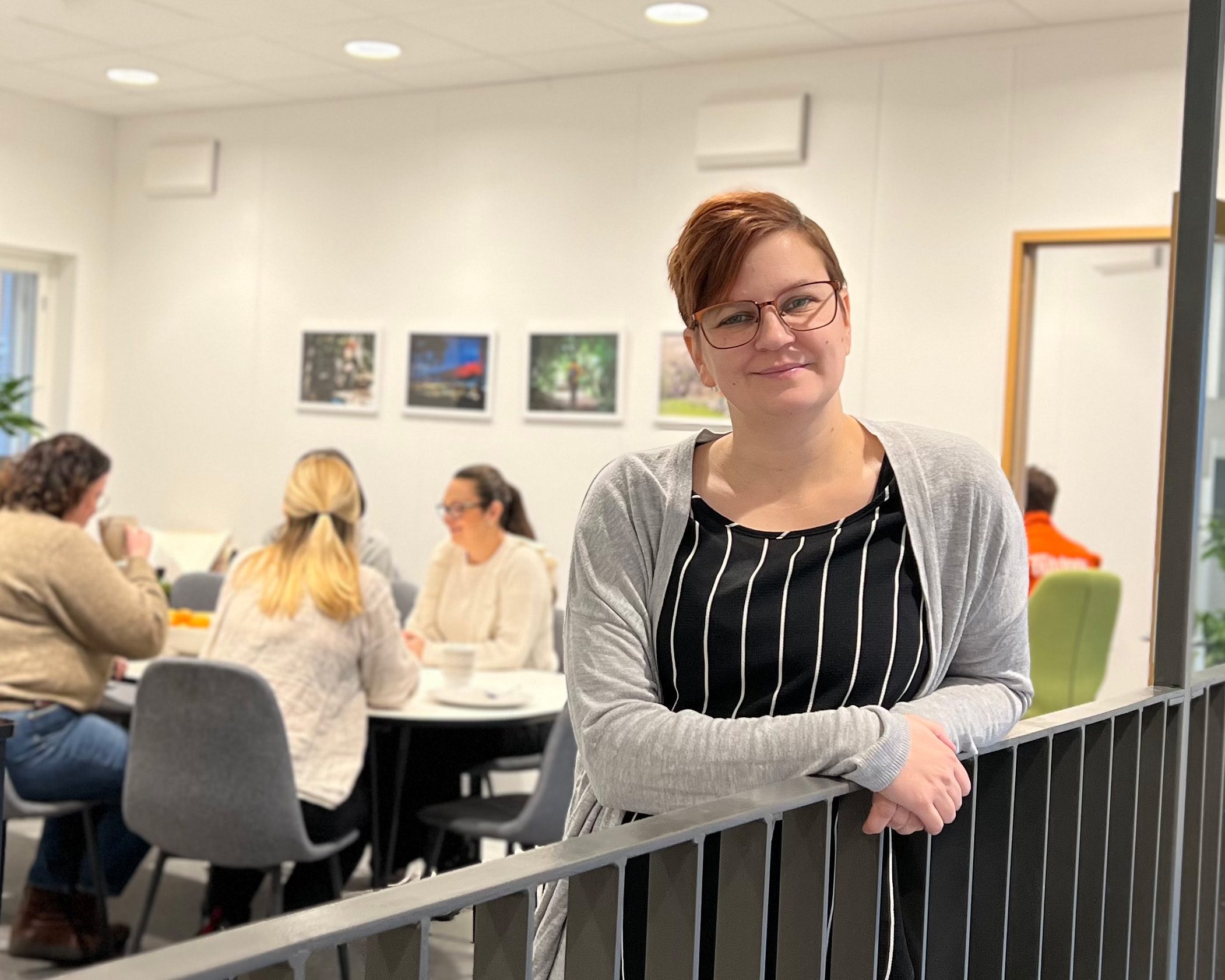 A woman with short red hair stands by a railing indoors. Behind her, people are eating lunch in a canteen in an office in Skövde.