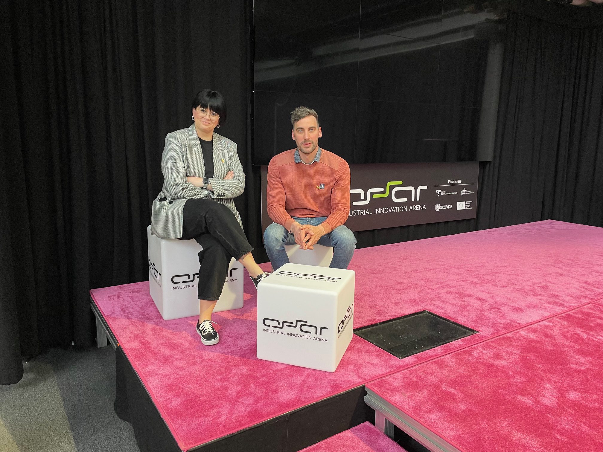 A man and a woman sit on a stage with a pink carpet.
