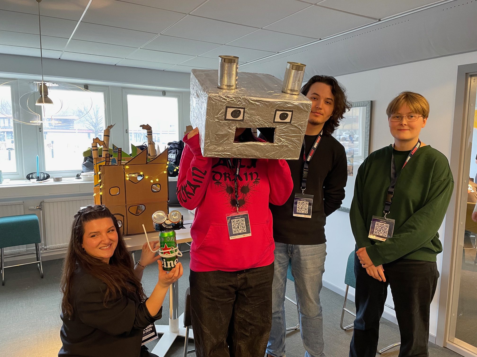 Fyra personer står framför en modell av en fabrik i papp. En person har en mask på huvudet för att likna en robot.