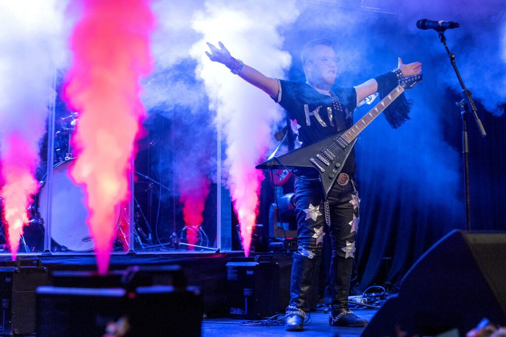 A man stands on stage with a guitarr around his neck and his arms in the air. In the background there are smoke canons going off.